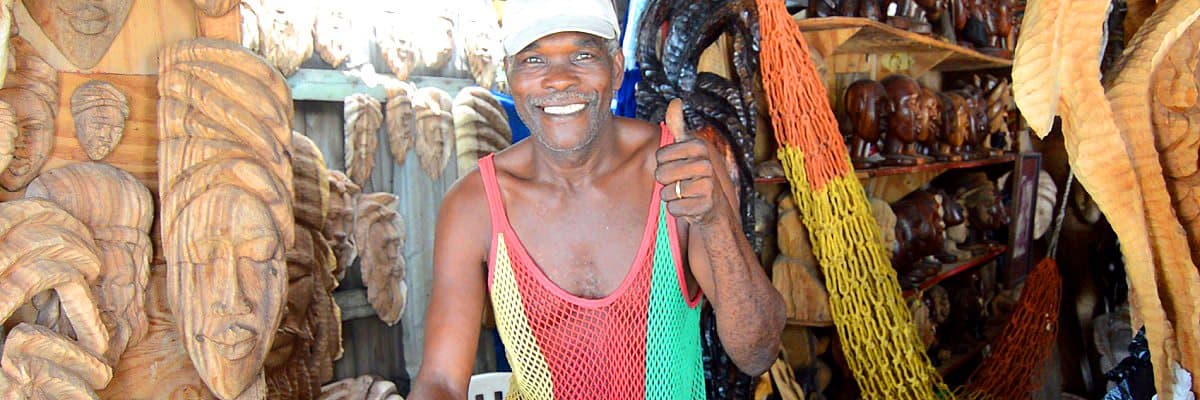 A smiling craft market vendor holding a woven bag, surrounded by carved wooden faces and figures