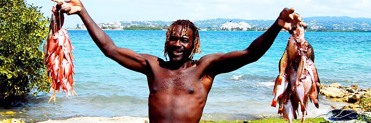 A fisherman on the shore at Montego Bay holding up two strings of red snapper, cruise ship behind him