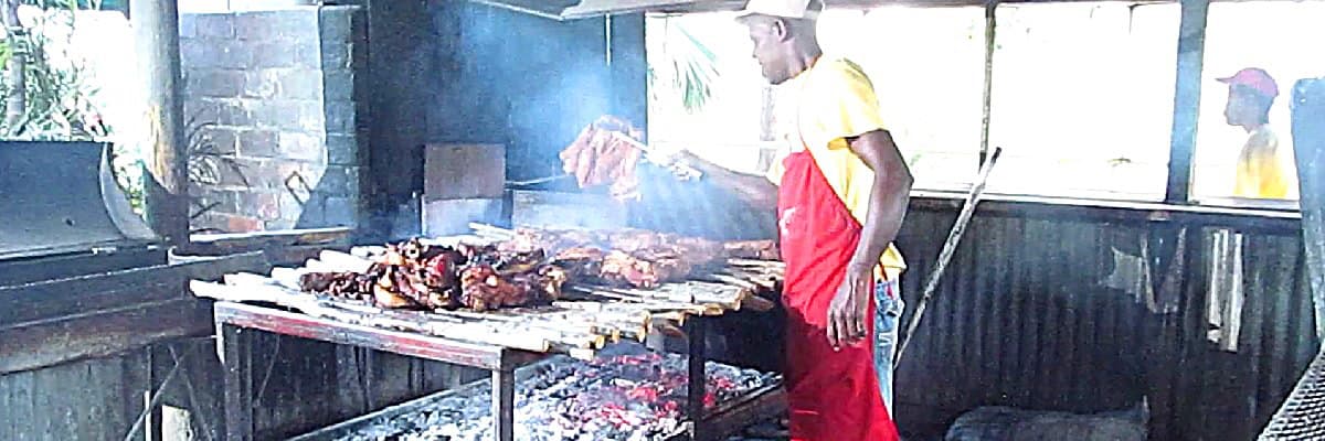 A cook in a yellow shirt turning jerk pork over a smoking open grill at a roadside jerk pit